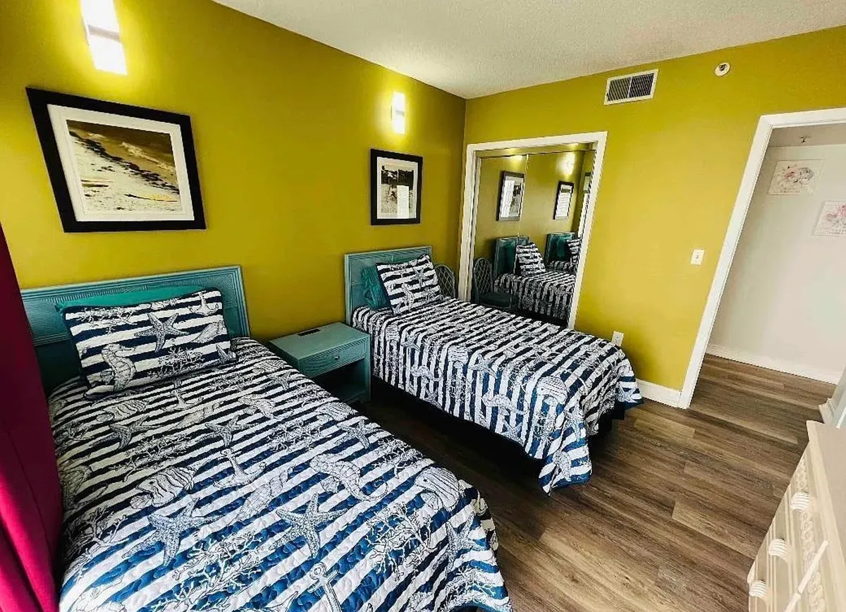 A bright bedroom featuring a bed with striped bedding, a colorful piece of wall art, and a nightstand with a lamp, all set in a space filled with morning light. The setup subtly suggests the presence of a relaxing area near the window, ideal for enjoying coffee while soaking in ocean views.