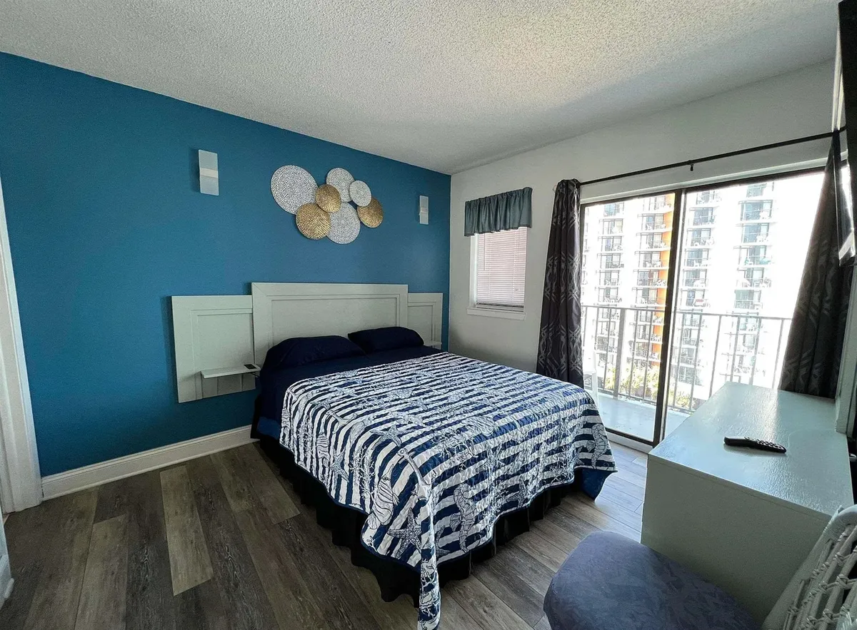 A tranquil primary bedroom with a comfortable bed, decorative wall art, soft linens, a balcony door, and window treatments that include room-darkening shades. The overall palette is calm and inviting, with natural light filtering softly into the space.