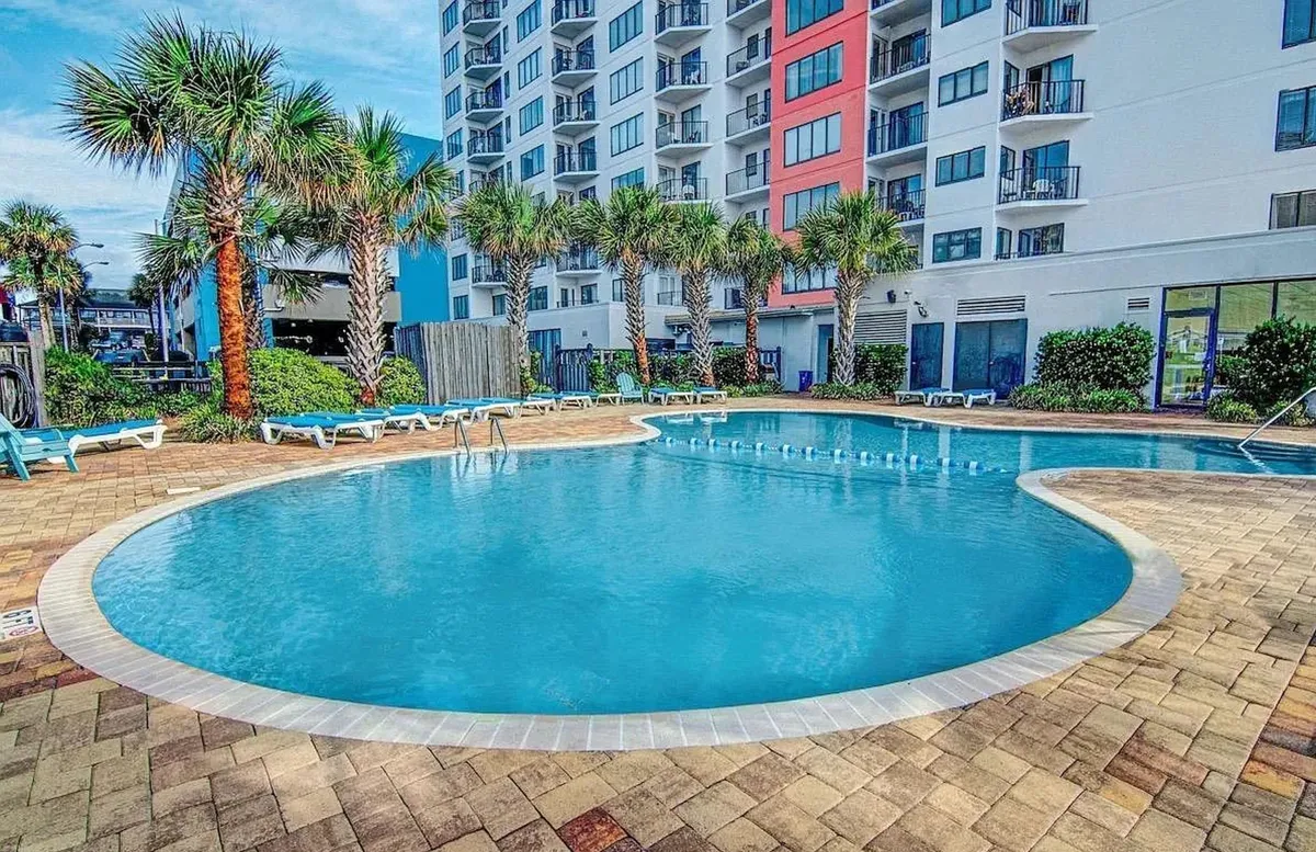 A sunlit exterior shot of the resort’s swimming pool area, framed by palm trees and lounge chairs, with the building’s facade visible in the background. The relaxed setting encourages leisure, sunbathing, and poolside gatherings.