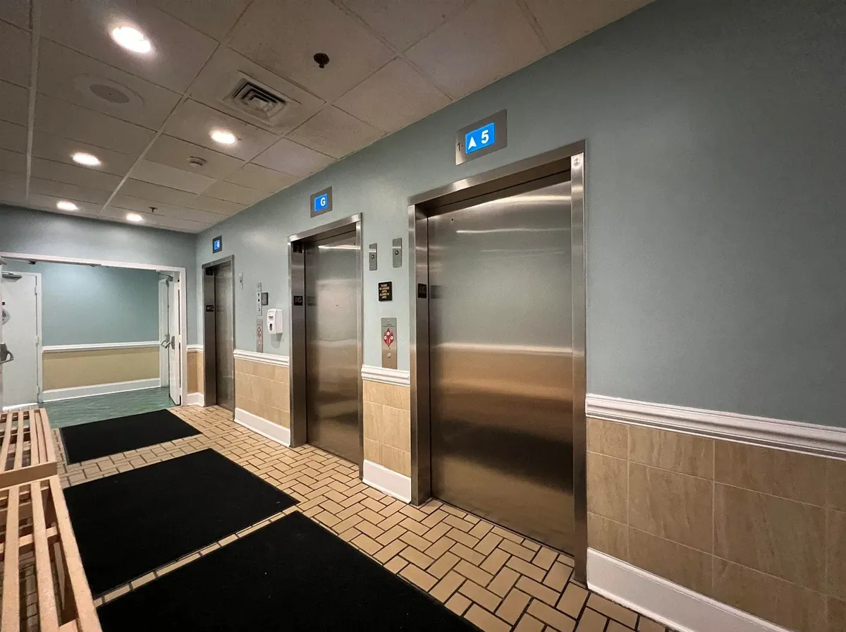 A bright, tiled corridor leading to elevators, with signage pointing toward beach access. The hallway is functional but hints at the convenience of getting from your condo directly to the sand, matching the narrative of quick and easy beach outings for couples.
