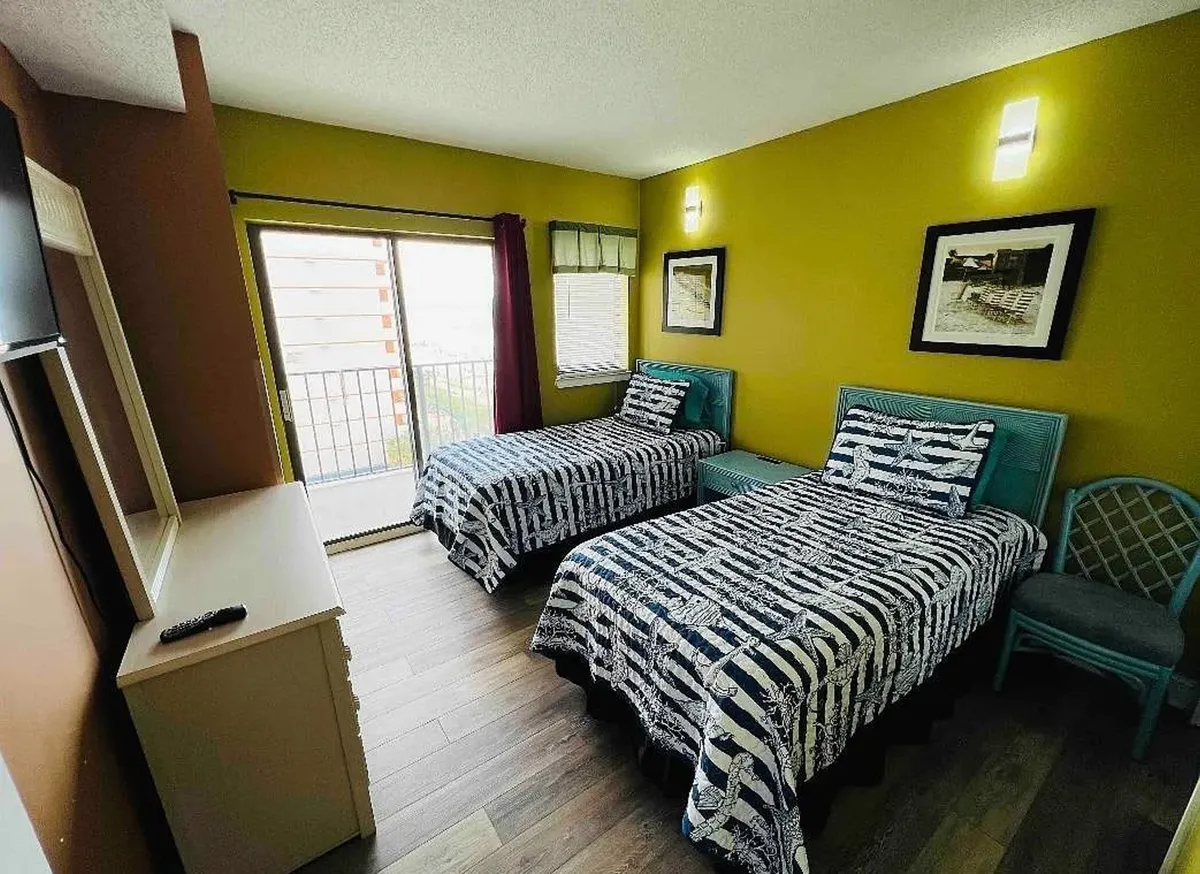 This image showcases a cozy interior space featuring two twin beds, vibrant wall art, and a glass door leading out to a private balcony. Natural light streams into the room, creating a cheerful ambiance and drawing attention to the inviting outdoor view.