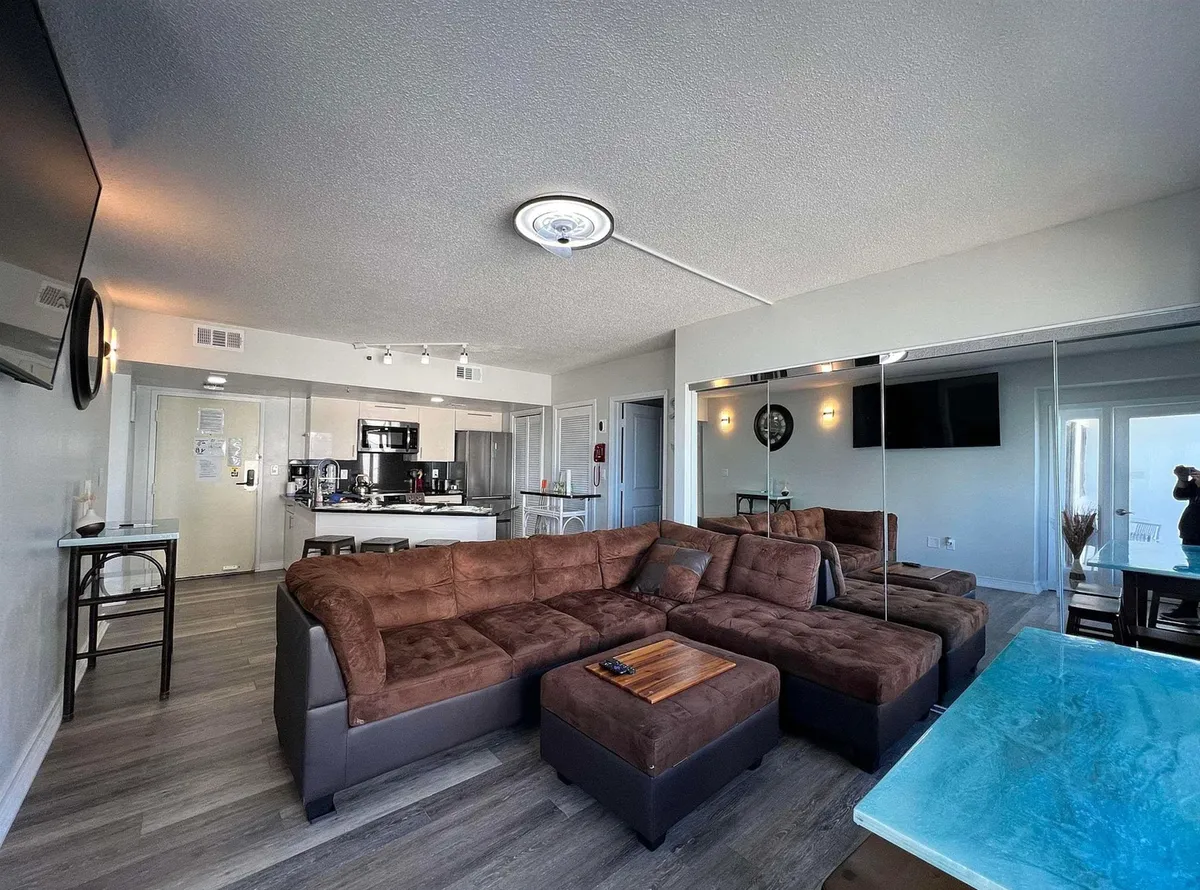 A contemporary living area with a brown sectional sofa, a glass-top table, a mirrored wall, a large TV, and a view into the kitchen. Subtle reflections enhance the sense of space and light, and the comfortable setup is perfect for streaming shows, entertaining, or lounging after a day on the beach.