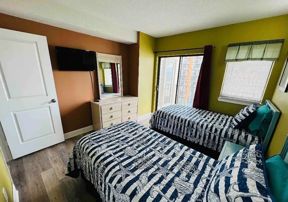 A cozy and cheerful second bedroom featuring two twin beds, a dresser with mirror, a TV on the wall, and a large window with a view. The room is bathed in natural light, making it adaptable for families or friends.