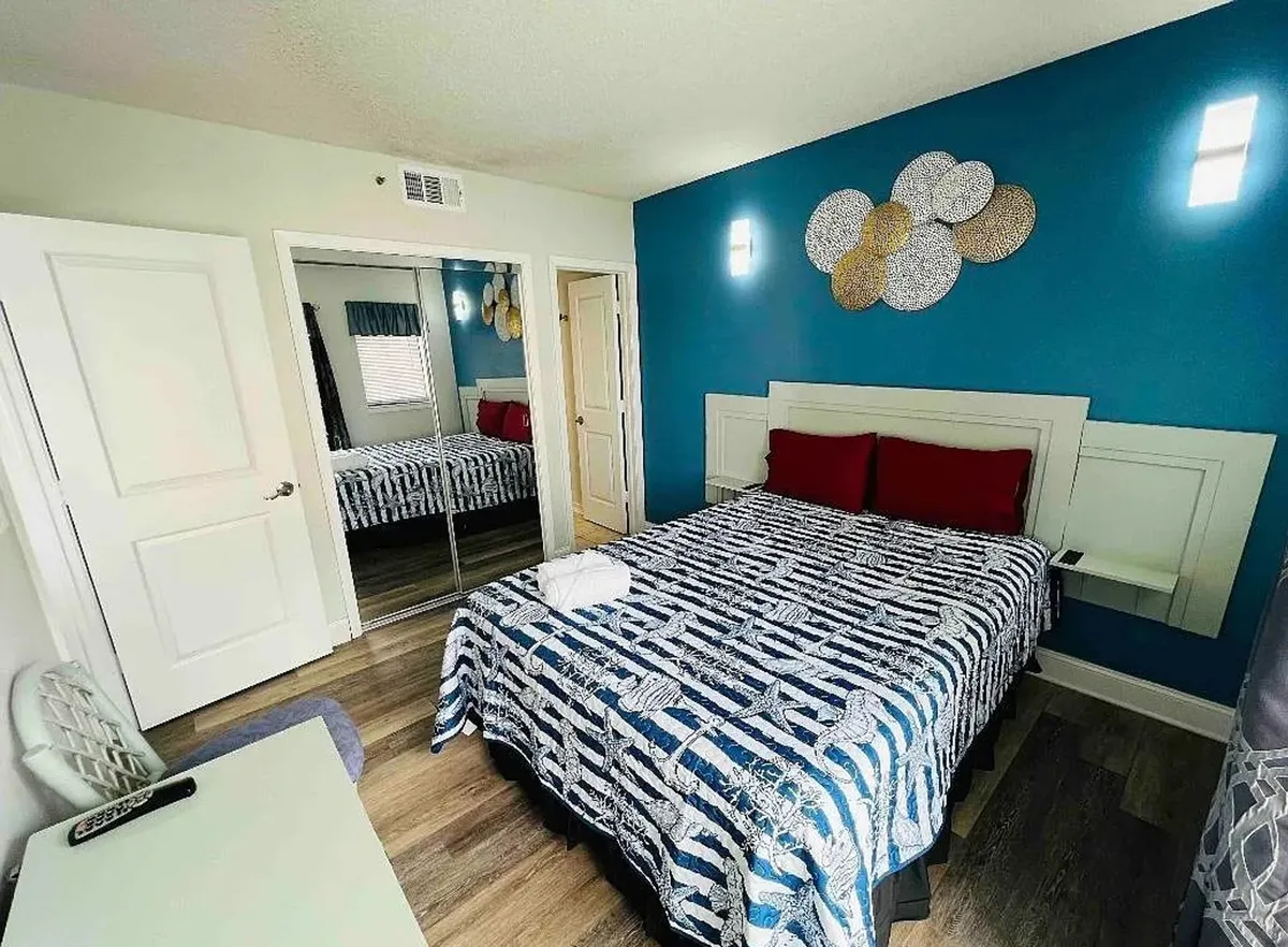 A cozy bedroom with a bed adorned with red pillows, decorative wall art, a mirror, and a desk area—bathed in natural light from a window, suggesting a welcoming environment for both productivity and downtime.